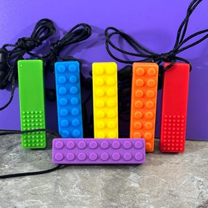 adult chewelry chew necklace - looks like a building brick block on the front and soothing bumps on the back. has a safety break away necklace. colors are: Red, orange. yellow, blue. green, purple