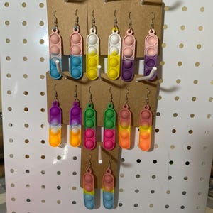 May include: A display of colorful pop it earrings. The earrings are made of silicone and have a variety of colors, including pink, blue, yellow, green, and purple. The earrings are displayed on a white pegboard.