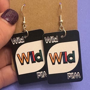 May include: A pair of Wild card earrings. Each earring features a black rectangular design with the word "Wild" at the top and the word "Wild" in colorful letters in the center. The earrings have silver-colored hooks.