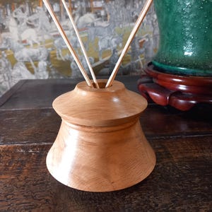 May include: A light brown wooden essential oil diffuser with three reeds. The diffuser has a rounded shape with a wider base and a narrower top. The reeds are thin and light-coloured, extending upwards from the diffuser.