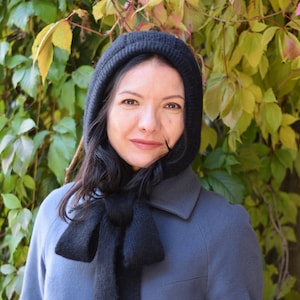 May include: A woman wearing a black hooded scarf and a dark blue coat. The scarf is tied in a large bow at the neck. The background features green and yellow foliage.