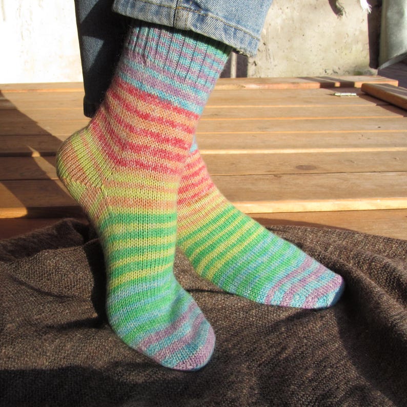 May include: A pair of colorful, striped socks in shades of green, yellow, pink, and blue. The socks are knitted and appear to be worn. The background shows wooden planks and a brown blanket.