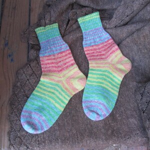 May include: A pair of hand-knitted socks with a rainbow color scheme. The socks feature horizontal stripes of blue, green, yellow, pink, and purple. The socks are displayed on a brown, textured surface.