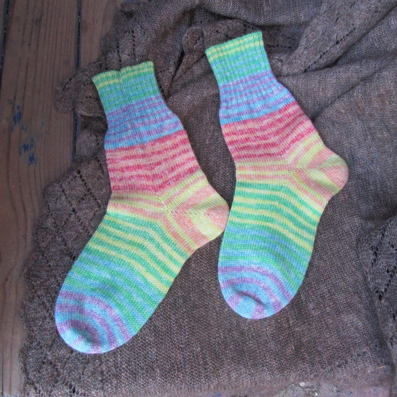May include: A pair of hand-knitted socks with a rainbow color scheme. The socks feature horizontal stripes of blue, green, yellow, pink, and purple. The socks are displayed on a brown, textured surface.