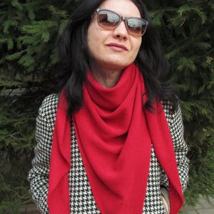 Puede incluir: Una persona con una bufanda roja brillante y gafas de sol. La bufanda está drapeada alrededor del cuello y los hombros. La chaqueta tiene un estampado de pata de gallo en blanco y negro.