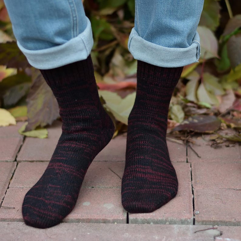 K&ouml;nnte beinhalten: Ein Paar schwarze und rote Socken mit einem melierten Muster. Die Socken werden von einer Person getragen, die eine blaue Jeans tr&auml;gt.
