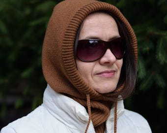 May include: A brown knitted balaclava with a drawstring closure. The balaclava is worn over a white jacket. The person in the image is wearing dark sunglasses. The background is a blurred green.