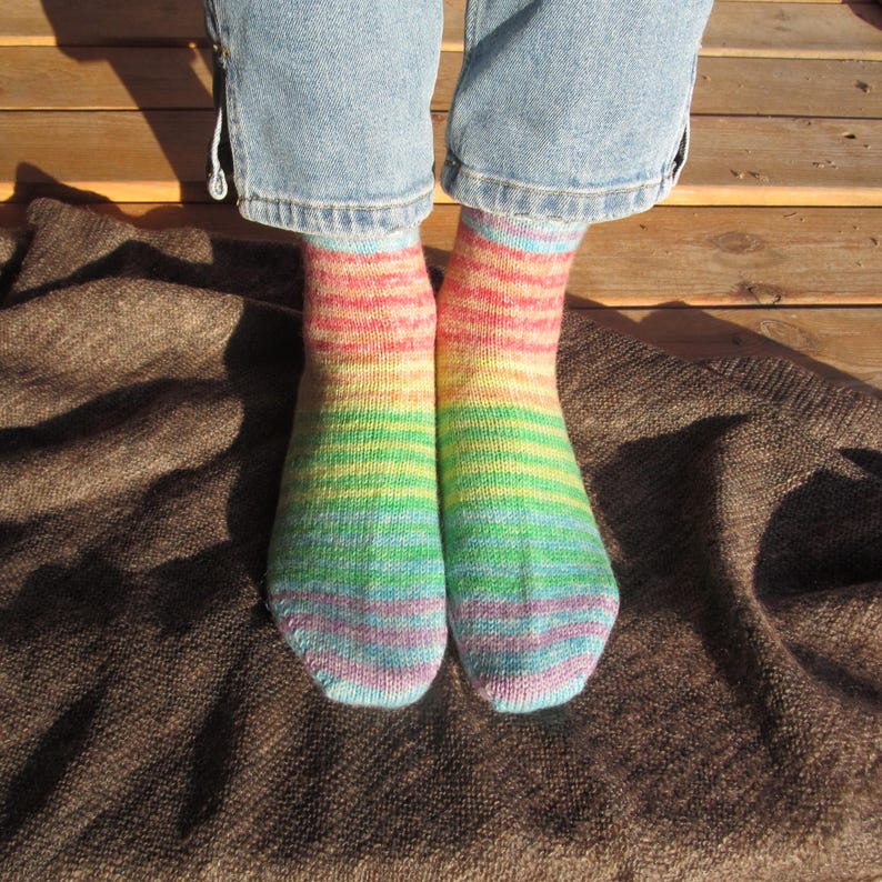 May include: A pair of colorful, hand-knitted socks with horizontal stripes in rainbow hues. The socks are worn with blue jeans and rest on a brown blanket. The socks feature shades of red, orange, yellow, green, blue, and purple.