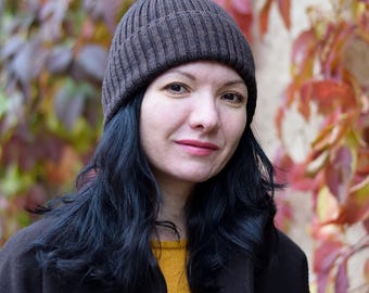 May include: A woman wearing a dark brown ribbed knit beanie, a dark brown coat, and a yellow sweater. Her dark hair frames her face, and she is smiling slightly. The background features colorful autumn foliage.