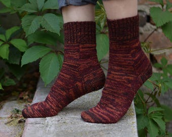 Könnte beinhalten: Ein Paar handgestrickte Socken in Burgunder-, Braun- und Orangetönen. Die Socken haben ein geripptes Bündchen und ein strukturiertes Muster am Bein und Fuß. Die Socken werden mit einer blauen Jeans getragen, vor dem Hintergrund von grünem Laub und einer Backsteinmauer.