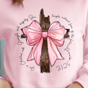 May include: A light pink sweatshirt featuring a graphic of a wooden cross adorned with a large pink bow. The design includes script that reads "Clothed with strength and dignity. She laughs without fear of the future. Proverbs 31:25."