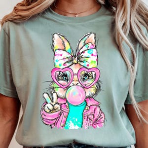 May include: A light green t-shirt featuring a cartoon bunny wearing heart-shaped sunglasses and a pink jacket, blowing a bubblegum bubble. The bunny has a colorful bow and is giving a peace sign.