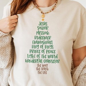 Könnte beinhalten: Beiges T-Shirt mit einem Weihnachtsbaum-Design aus religiösen Titeln in Grün und Braun. Der Text lautet: "Jesus, Retter, Messias, Erlöser, Immanuel, König der Könige, Friedefürst, Licht der Welt, Wunderbarer Ratgeber, Der Weg, Die Wahrheit, Das Leben."