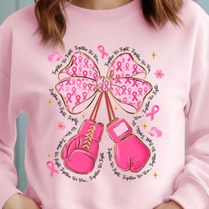 May include: Pink sweatshirt featuring a large pink ribbon with pink ribbon accents and boxing gloves. The words "Together We Fight" and "Together We Win" are written around the design. The sweatshirt is a light pink color.