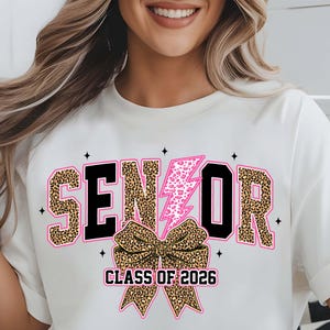 Może przedstawiać: Kremowy t-shirt z napisem "SENIOR" w panterkę i czarnymi literami. Różowa błyskawica z akcentami w panterkę akcentuje literę "O". Poniżej znajduje się kokarda w panterkę i napis "CLASS OF 2026".
