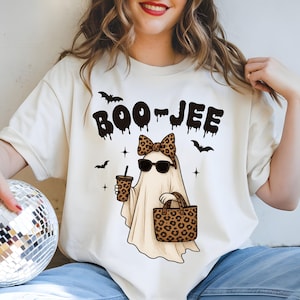 May include: Cream-colored t-shirt with a Halloween-themed graphic. The design features a cartoon ghost wearing sunglasses and a leopard print bow, holding a coffee cup and a leopard print tote bag. The text "BOO-JEE" is above the ghost, with bat and star accents.