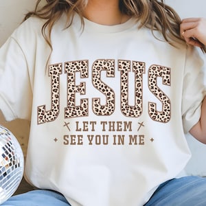 May include: Cream-coloured t-shirt featuring the word "JESUS" in large leopard print letters. Below the word "JESUS" are the words "LET THEM SEE YOU IN ME" in a brown font. The shirt has short sleeves.
