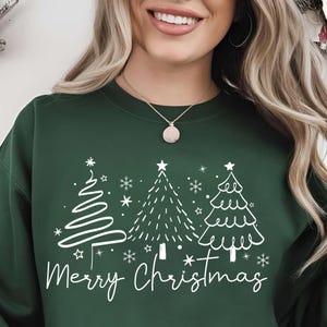 Puede incluir: Sudadera verde oscuro con un gráfico blanco de tres árboles de Navidad y copos de nieve. Las palabras "Merry Christmas" están escritas en cursiva debajo de los árboles. Una prenda festiva para la temporada navideña.