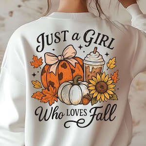 May include: White sweatshirt with a fall-themed graphic. The design includes an orange pumpkin with a bow, a white pumpkin, a coffee drink, a sunflower, and fall leaves. Text reads "Just a Girl Who Loves Fall."