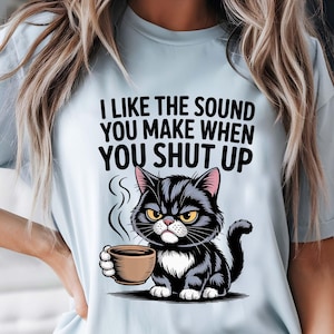 Puede incluir: Camiseta azul claro con un gato negro de dibujos animados sosteniendo una taza de café. El gato tiene ojos amarillos y una expresión enfadada. La camiseta tiene el texto "I LIKE THE SOUND YOU MAKE WHEN YOU SHUT UP" en negro.