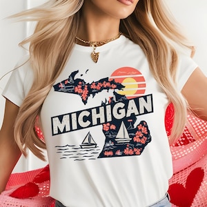 Puede incluir: Camiseta blanca con un gráfico del estado de Michigan, con una puesta de sol, veleros y flores. El texto "MICHIGAN" está impreso en letras azules oscuras en negrita.