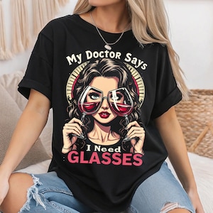 Può includere: Maglietta nera con una grafica di una donna che indossa occhiali fatti di bicchieri di vino. Il testo sulla maglietta recita "My Doctor Says I Need Glasses".
