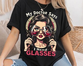 Op de afbeelding: Zwart T-shirt met een afbeelding van een vrouw die een bril draagt ​​gemaakt van wijnglazen. De tekst op het shirt luidt "My Doctor Says I Need Glasses".