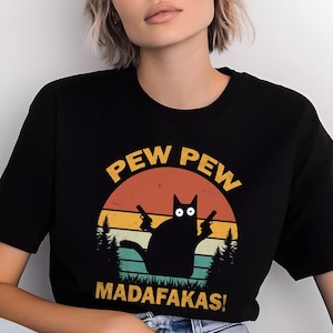 May include: Black t-shirt featuring a graphic of a black cat holding two guns, with the words "PEW PEW MADAFAKAS!" in yellow. The design includes a sunset and trees.