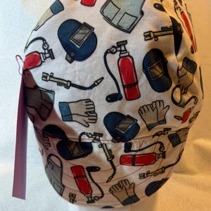 May include: A white fabric cap featuring a repeating pattern of welding equipment including red fire extinguishers, blue welding helmets, tan work gloves, grey aprons, and yellow welding torches. A small pink tag is attached to the side.