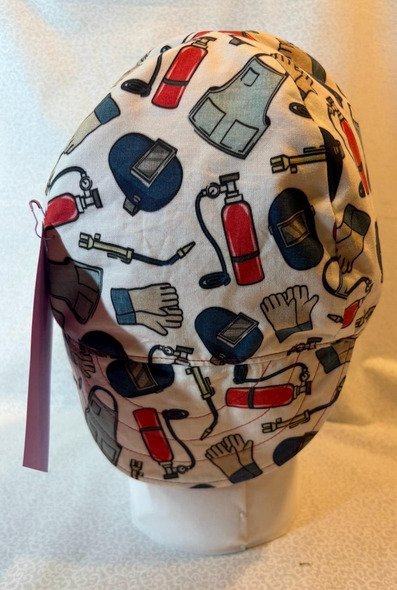 May include: A white fabric cap featuring a repeating pattern of welding equipment including red fire extinguishers, blue welding helmets, tan work gloves, grey aprons, and yellow welding torches. A small pink tag is attached to the side.
