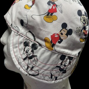 May include: A white welding cap featuring a repeating pattern of Mickey Mouse in various poses. The character is depicted in black, white, red, and yellow colors. The cap has a rounded crown and a curved brim with visible red stitching details.