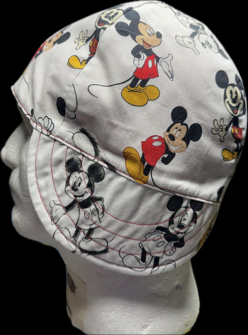May include: A white welding cap featuring a repeating pattern of Mickey Mouse in various poses. The character is depicted in black, white, red, and yellow colors. The cap has a rounded crown and a curved brim with visible red stitching details.