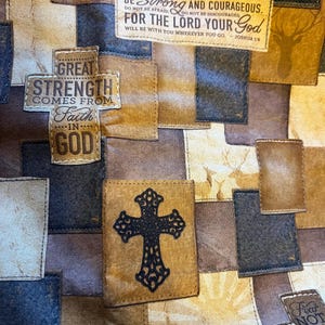 May include: A fabric quilt featuring a patchwork design in shades of brown, tan, and navy blue. The pattern includes printed text such as Be Strong and Courageous, For the Lord Your God, and Great Strength Comes from Faith in God, alongside a black cross emblem and deer silhouettes.