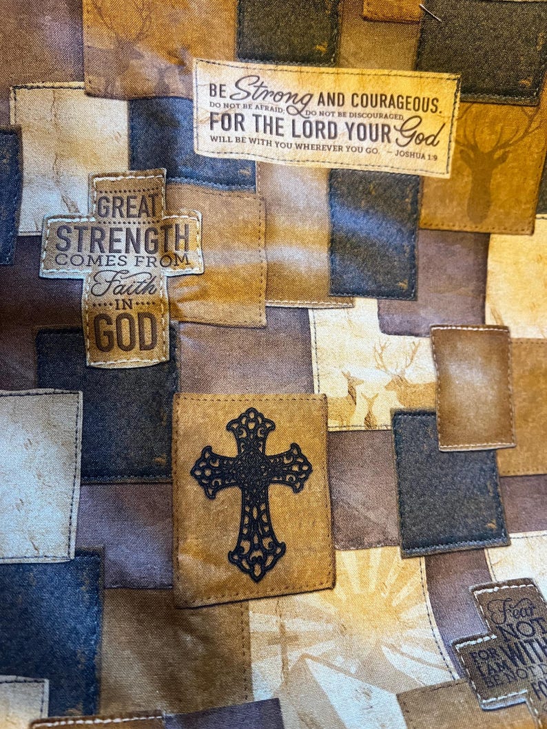 May include: A fabric quilt featuring a patchwork design in shades of brown, tan, and navy blue. The pattern includes printed text such as Be Strong and Courageous, For the Lord Your God, and Great Strength Comes from Faith in God, alongside a black cross emblem and deer silhouettes.