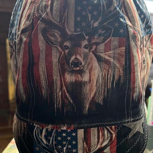 May include: A fabric welding cap featuring a repeating print of a deer head centered over a stylized American flag pattern with red, white, and blue colors. The cap has a rounded crown and a short, stitched brim, constructed with visible seams.