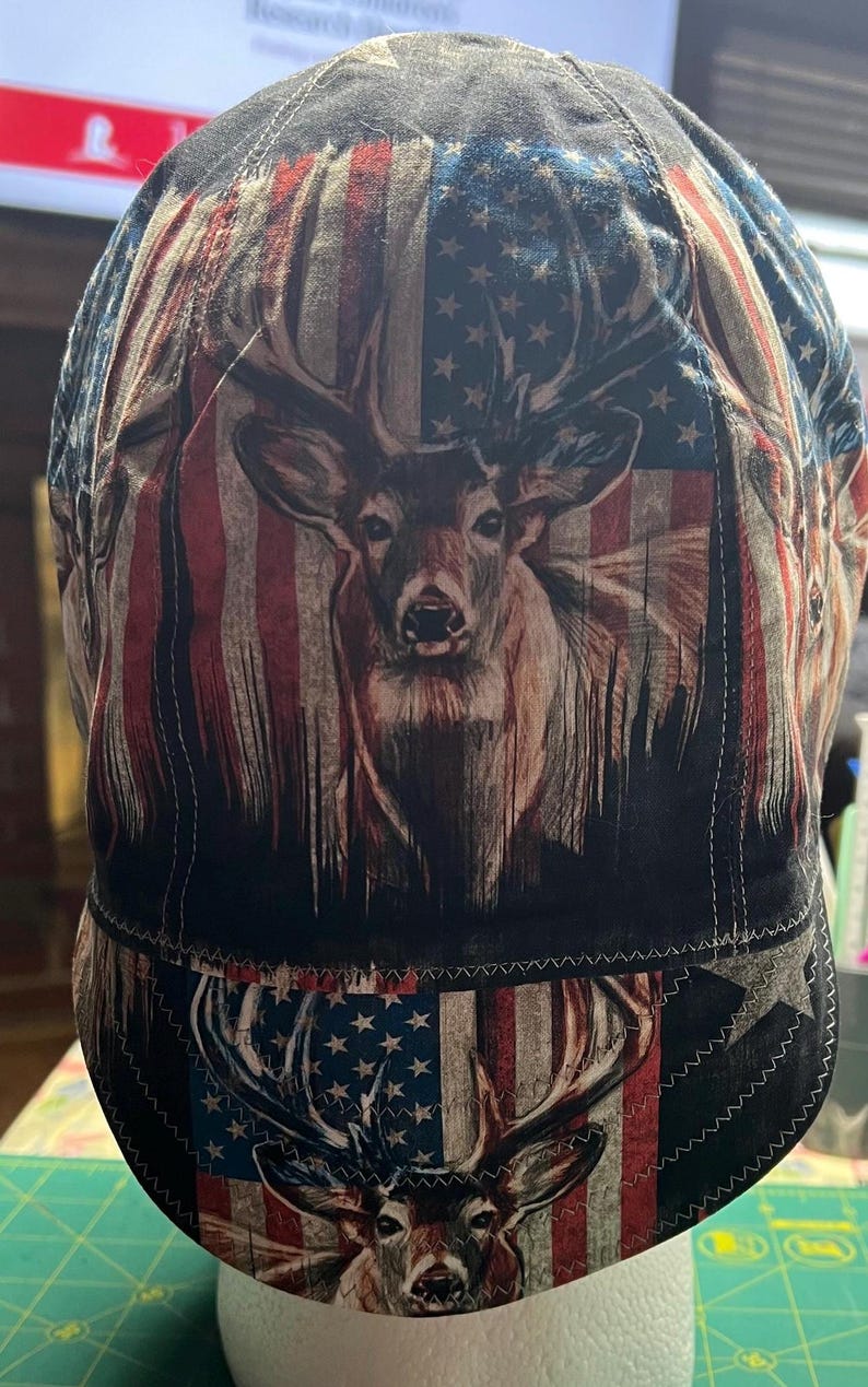 May include: A fabric welding cap featuring a repeating print of a deer head centered over a stylized American flag pattern with red, white, and blue colors. The cap has a rounded crown and a short, stitched brim, constructed with visible seams.