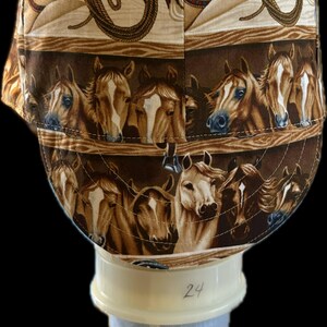May include: A fabric cap featuring a repeating pattern of brown and white horses, cowboy hats, and coiled lassos on a tan background. The cap has a rounded crown and a short brim, with visible stitching throughout the design and the number 24.