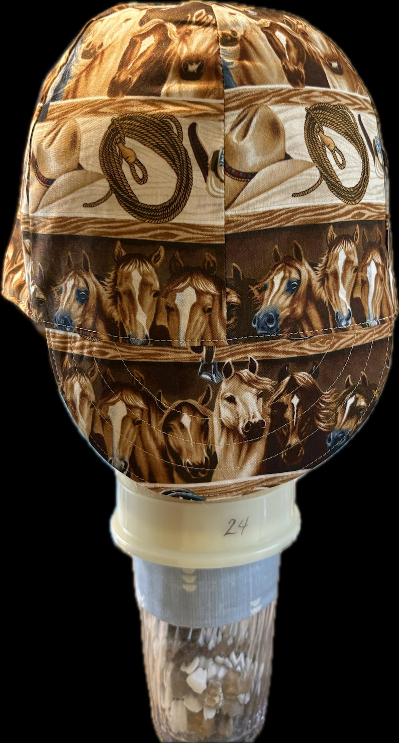 May include: A fabric cap featuring a repeating pattern of brown and white horses, cowboy hats, and coiled lassos on a tan background. The cap has a rounded crown and a short brim, with visible stitching throughout the design and the number 24.