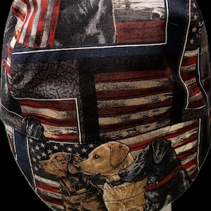 May include: A scrub cap featuring a repeating pattern of American flags and various dog breeds, including a brown dog, a yellow Labrador, and a black dog. The fabric design incorporates red, white, and blue colors with detailed illustrations of dogs.