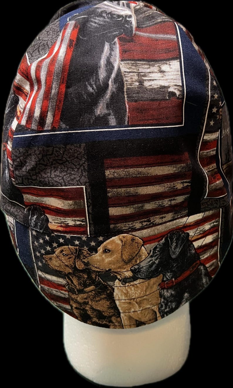 May include: A scrub cap featuring a repeating pattern of American flags and various dog breeds, including a brown dog, a yellow Labrador, and a black dog. The fabric design incorporates red, white, and blue colors with detailed illustrations of dogs.