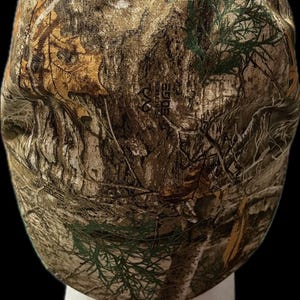 May include: A baseball cap featuring a RealTree Edge camouflage pattern in shades of brown, tan, green, and white. The fabric displays a detailed print of tree bark, branches, and leaves. The RealTree Edge logo is visible in the center of the cap.