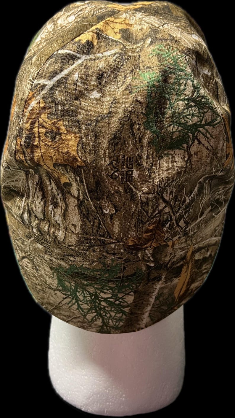 May include: A baseball cap featuring a RealTree Edge camouflage pattern in shades of brown, tan, green, and white. The fabric displays a detailed print of tree bark, branches, and leaves. The RealTree Edge logo is visible in the center of the cap.
