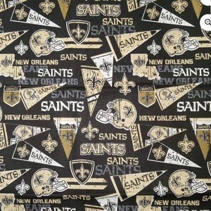 May include: A black fabric featuring a repeating pattern of New Orleans Saints football team graphics. The design includes gold and white football helmets, pennants with the text SAINTS, fleur-de-lis symbols, and the text NEW ORLEANS.