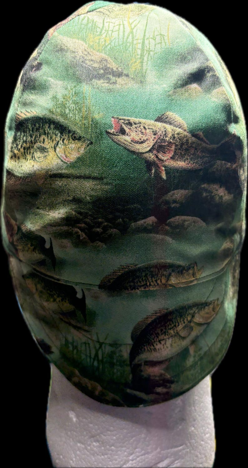 May include: A baseball cap featuring an all-over print of various fish swimming in a green underwater environment with rocks and aquatic plants. The design displays multiple fish species in shades of brown, green, and yellow against a green background.