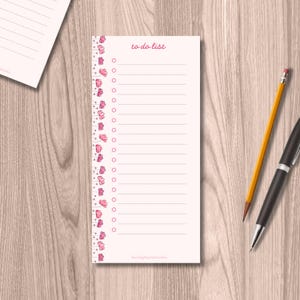 May include: A pale pink to-do list notepad with a floral border of pink butterflies. The words "to do list" are written in pink script at the top. A yellow pencil and a black pen are on the wooden surface.