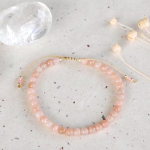 May include: A light pink beaded bracelet with an adjustable knot closure. The bracelet features small, round, pale pink beads. Two polished stones and dried flowers are also visible in the image.