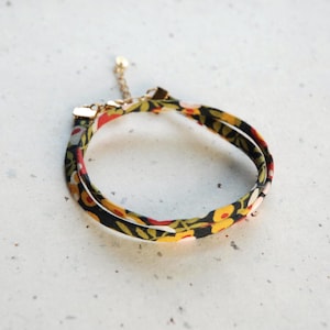May include: A double-wrap bracelet featuring a black fabric band with a colourful floral pattern in shades of yellow, red, and green. The bracelet includes gold-toned metal end caps and a delicate gold-toned chain extender with a small circular charm.