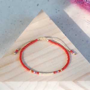 May include: A delicate bracelet featuring a continuous strand of small red seed beads, accented with a central section of metallic gold beads and small purple beads. The bracelet includes an adjustable sliding knot closure with matching bead ends.