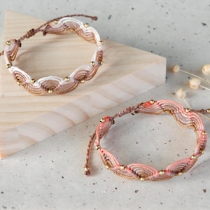 May include: Two macrame bracelets with a wave-like design. One bracelet is in shades of brown and cream, the other in pink and gold. Both have gold beads and brown cords. They are displayed on a wooden triangle and a speckled surface.