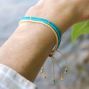 May include: A delicate beaded bracelet with turquoise and cream-coloured beads. The bracelet has gold accents and adjustable cords. The bracelet is shown on an arm, with a blurred background of water and foliage.